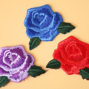 Könnte beinhalten: Drei gestickte Bügelbilder in Form von Rosen. Die Rosen sind blau, lila und rot.