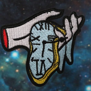 May include: An embroidered patch of a hand holding a clock with Roman numerals. The clock is blue and yellow, and the hand is white with red accents.
