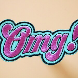 May include: A turquoise and pink embroidered patch with the word "Omg!" in a stylized font.