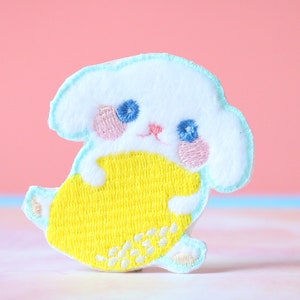 Może przedstawiać: Biała haftowana łatka z postacią kreskówkowego zwierzęcia z niebieskimi oczami, różowymi policzkami i żółtym ciałem. Zwierzę trzyma w łapkach żółty owoc.