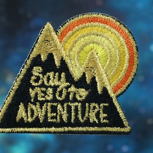 May include: A black triangular patch with gold embroidery depicting a mountain and a setting sun. The text "Soul Seekers Adventure" is embroidered in gold on the patch.