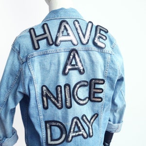 Puede incluir: Una chaqueta vaquera azul claro con las palabras "Have a Nice Day" escritas en lentejuelas plateadas en la espalda.