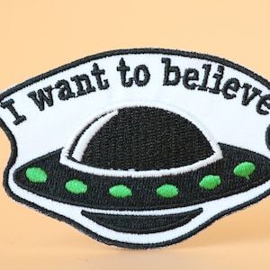 Może przedstawiać: Biała haftowana naszywka z czarnym UFO i tekstem "Chcę wierzyć".