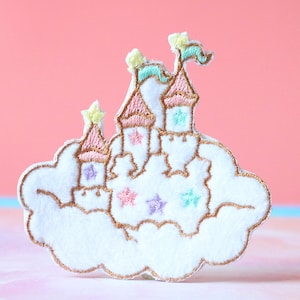 Puede incluir: Un parche bordado de fieltro blanco con un castillo de color rosa pastel y azul sobre una nube con costuras doradas. El castillo tiene una bandera y estrellas.
