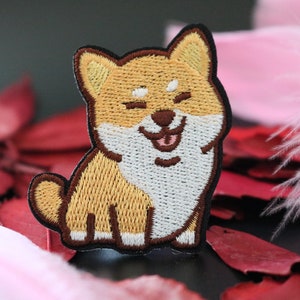 May include: Embroidered patch of a brown and white Shiba Inu dog with a happy expression. The dog is sitting with its tongue sticking out.