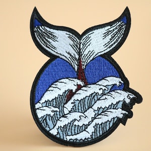 May include: Embroidered iron-on patch featuring a white whale tail emerging from blue water with white waves. The patch has a black outline.