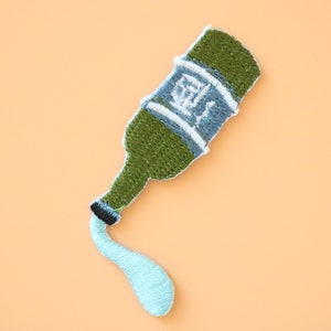 May include: Green embroidered patch of a bottle pouring a blue liquid. The bottle has white text on it.