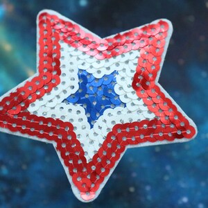 Könnte beinhalten: Ein sternförmiger Patch in Rot, Weiß und Blau aus Pailletten. Der Stern ist mit roten Pailletten umrandet, hat eine weiße Mitte und einen blauen Stern in der Mitte.