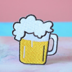 Könnte beinhalten: Ein gestickter Bügelaufkleber mit einem Bierkrug mit weißem Schaumkopf und gelbem Bier.