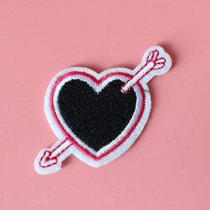 Puede incluir: Un parche blanco en forma de corazón con un contorno rojo y un corazón negro en el interior. Una flecha con una punta roja y plumas blancas atraviesa el corazón.