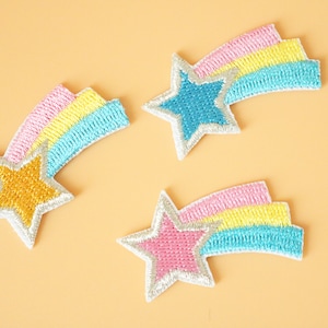 May include: Three embroidered iron-on patches in the shape of shooting stars with rainbow tails. The stars are yellow, blue, and pink, and the tails are pink, yellow, and blue.