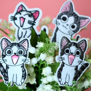 May include: Four embroidered iron-on patches featuring a cartoon cat with black and white fur and pink ears. The cat is smiling and has large eyes. The patches are on a background of white flowers.