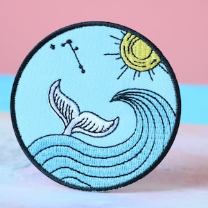 May include: Embroidered patch featuring a whale tail emerging from blue waves under a yellow sun. A constellation is above the whale tail. The patch has a black border and is on a light blue background.