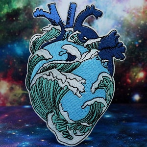 Puede incluir: Un parche bordado de un corazón azul con olas rompiendo sobre él. El corazón está contorneado en negro y las olas son en tonos de azul y verde.