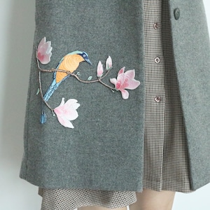 May include: A gray wool coat with a bird and flower embroidery on the front. The embroidery is in shades of blue, yellow, and pink. The coat has a checkered pattern on the inside.
