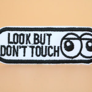 以下が含まれることがあります： 黒い文字で「Look But Don't Touch」と書いてある白い刺繍パッチと、黒い漫画の目が2つある。