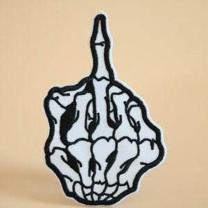 May include: A white embroidered patch of a skeleton hand giving the middle finger. The patch is outlined in black thread.