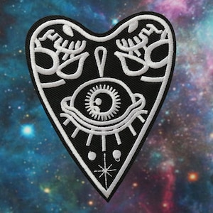 May include: Black and white embroidered patch in the shape of a heart with a single eye inside. The eye is surrounded by a white circle and has a black pupil. There are two white dots and a star at the bottom of the patch.