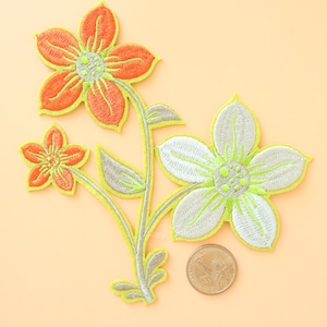 May include: Three embroidered flower patches in orange, white, and green. The flowers are arranged in a cluster with stems and leaves. The patches are approximately the size of a US quarter.
