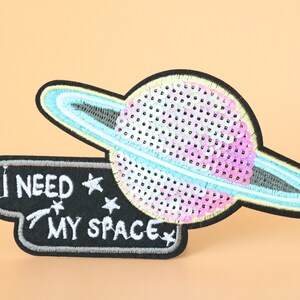 May include: A black and grey embroidered patch with a pink and blue sequin planet and the text "I NEED MY SPACE" with stars.