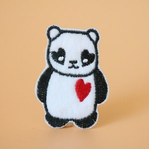 May include: A black and white embroidered panda bear with a red heart on its chest. The panda is standing on a light orange background.