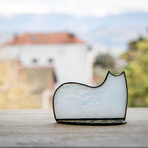 May include: A stained glass cat figurine featuring a white and pale blue marbled pattern with a smooth, curved silhouette. The piece is outlined in a dark, thin metal solder frame that defines the shape of the cat's back, ears, and base.