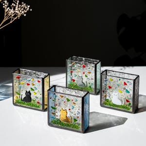 Könnte beinhalten: Vier quadratische Kerzenhalter aus Glas mit farbenfrohen Glasmalereien. Jeder Halter zeigt eine Katzenillustration in Schwarz, Grau, Weiß und Orange. Die Halter haben eine grüne Grasbasis und farbige Akzente.