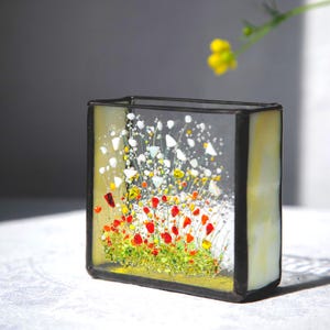 Könnte beinhalten: Eine rechteckige Glasvase mit schwarzem Metallrahmen. Die Vase zeigt ein farbenfrohes Blumendesign mit roten, weißen, gelben und grünen Glaselementen. Das Glas hat einen hellgelben Farbton, und das Gesamtdesign ist künstlerisch und dekorativ.