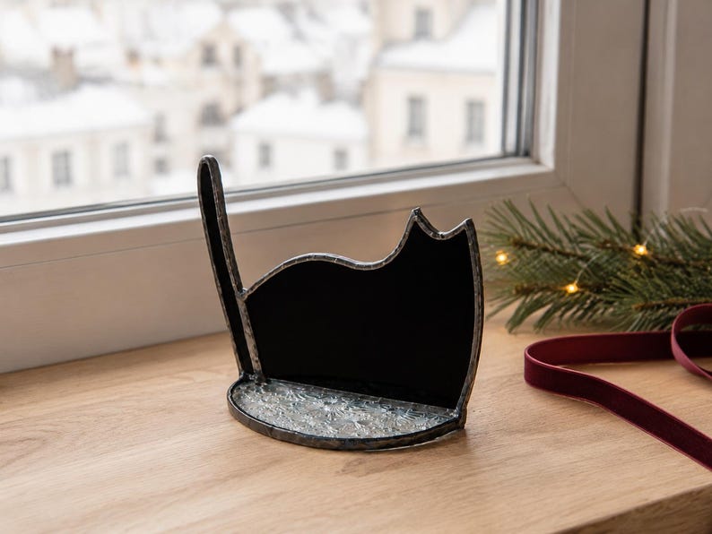 May include: A black stained glass cat silhouette with a raised tail, featuring a textured, clear glass base. The cat figure is outlined in dark metal solder, creating a defined shape that stands upright on a flat surface.
