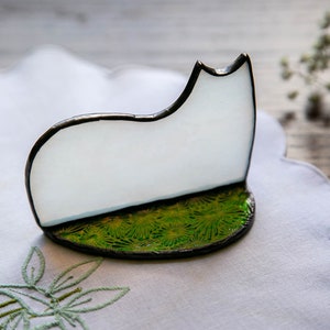 May include: A stained glass cat silhouette featuring a smooth, opaque white body with a dark metal solder frame. The cat stands on a circular base decorated with a vibrant green, textured pattern resembling grass, also edged in dark metal solder.