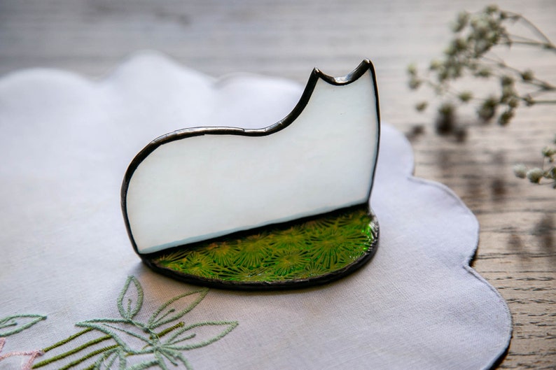 May include: A stained glass cat silhouette featuring a smooth, opaque white body with a dark metal solder frame. The cat stands on a circular base decorated with a vibrant green, textured pattern resembling grass, also edged in dark metal solder.