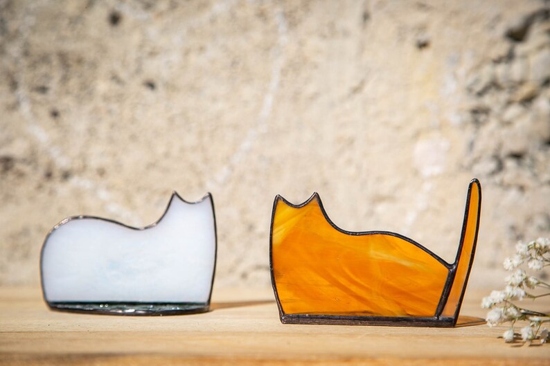 May include: Two stained glass cat figurines with dark metal soldered edges. One cat is made of opaque white glass with a smooth, rounded back. The other cat is made of translucent orange glass with a visible tail pointing upward.