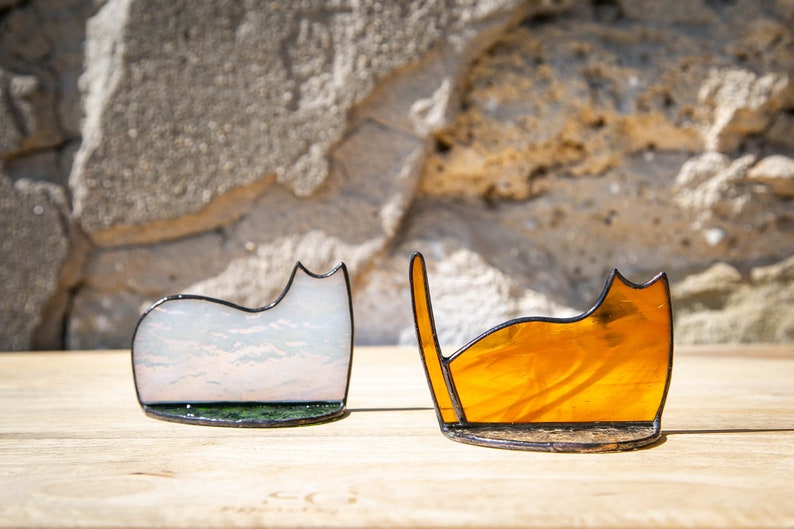 May include: Two stained glass cat figurines with black metal soldered edges. One cat is made of translucent white glass with subtle cloud-like patterns, and the other is made of vibrant amber-colored glass with a raised tail silhouette.