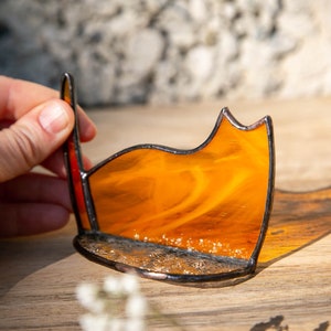 May include: A stained glass cat silhouette in vibrant orange with a marbled texture, featuring dark soldered metal edges and a textured base. The piece is crafted with a curved back and a pointed ear, showcasing translucent glass that casts a warm shadow.