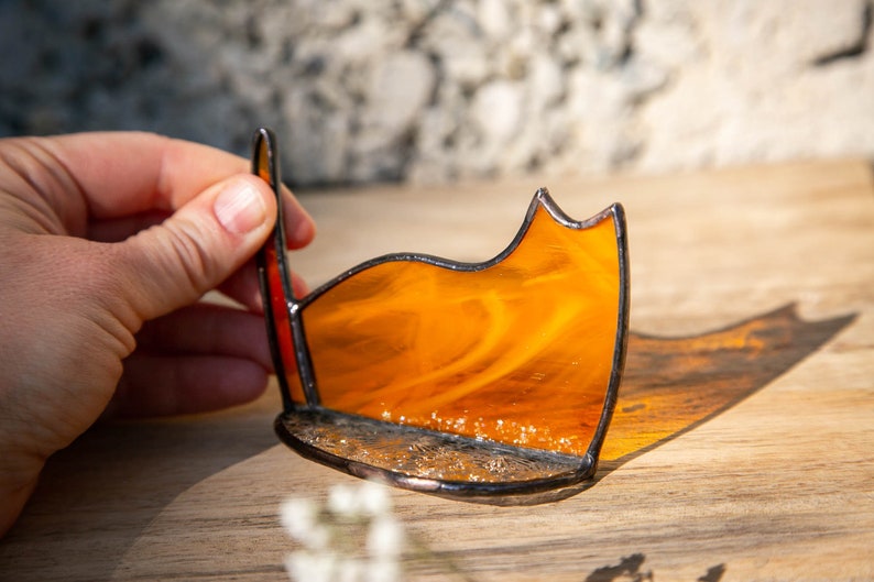 May include: A stained glass cat silhouette in vibrant orange with a marbled texture, featuring dark soldered metal edges and a textured base. The piece is crafted with a curved back and a pointed ear, showcasing translucent glass that casts a warm shadow.