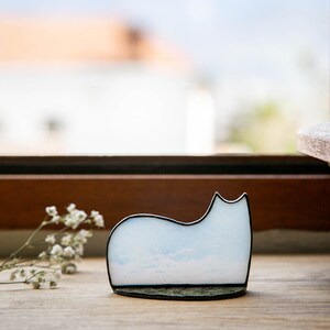 May include: A cat-shaped decorative object made of light blue and white glass with a cloudy pattern, framed in a thin black metal border. The piece features a smooth, curved silhouette and sits on a flat, dark, textured base next to a small sprig.
