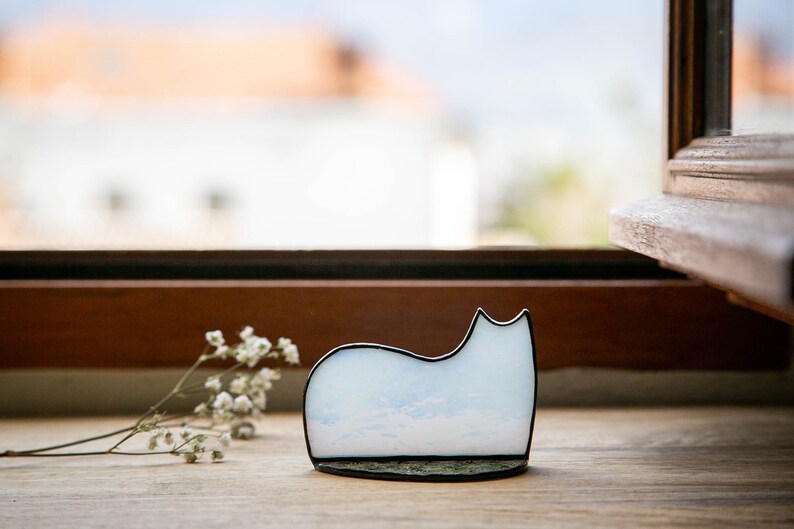 May include: A cat-shaped decorative object made of light blue and white glass with a cloudy pattern, framed in a thin black metal border. The piece features a smooth, curved silhouette and sits on a flat, dark, textured base next to a small sprig.