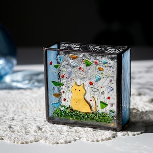 May include: A stained glass box with a yellow cat silhouette in a garden scene. The box has blue sides and is filled with colorful glass pieces. The cat is on a green base, with colorful flowers and glass accents.