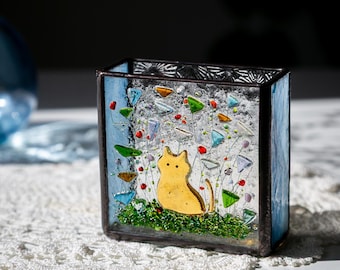 May include: A stained glass box with a yellow cat silhouette in a garden scene. The box has blue sides and is filled with colorful glass pieces. The cat is on a green base, with colorful flowers and glass accents.