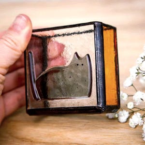 May include: A square, glass candle holder with a cat silhouette design. The holder has a metallic finish with a black border and amber-colored side. The cat is etched into the glass. The holder is held by a hand.