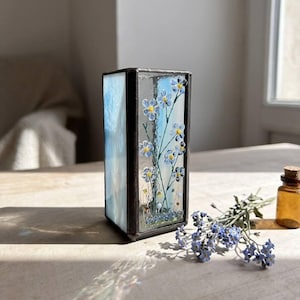 May include: A rectangular stained glass box with a black metal frame. The glass panels feature a blue sky design and delicate blue and yellow forget-me-not flowers. A small bottle and sprig of flowers are next to the box.