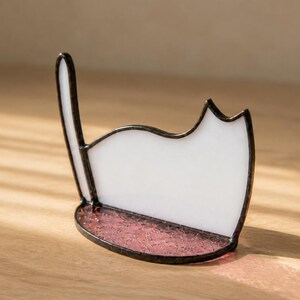 May include: A stained glass cat silhouette ornament featuring a white opaque body and a tall upright tail. The base is a semi-circular piece of textured, translucent pink glass. The entire structure is held together by dark metal solder lines.