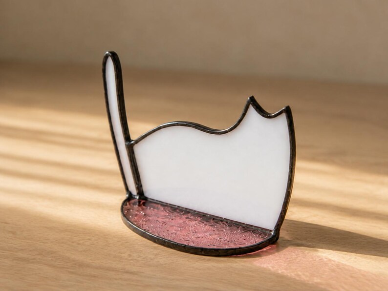 May include: A stained glass cat silhouette ornament featuring a white opaque body and a tall upright tail. The base is a semi-circular piece of textured, translucent pink glass. The entire structure is held together by dark metal solder lines.