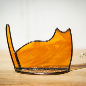 May include: A stained glass cat silhouette ornament crafted from translucent amber-colored glass with a marbled texture. The piece features dark metal solder lines outlining the cat's form, including a pointed ear and an upright tail, set on a base.