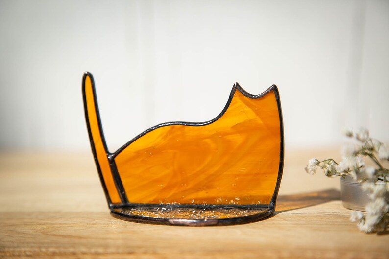 May include: A stained glass cat silhouette ornament crafted from translucent amber-colored glass with a marbled texture. The piece features dark metal solder lines outlining the cat's form, including a pointed ear and an upright tail, set on a base.