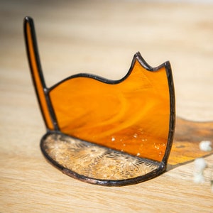 May include: A stained glass cat silhouette ornament featuring a translucent amber-colored body with a dark metal solder frame. The base is a semi-circular piece of clear textured glass with a starburst pattern, joined to the upright cat figure.