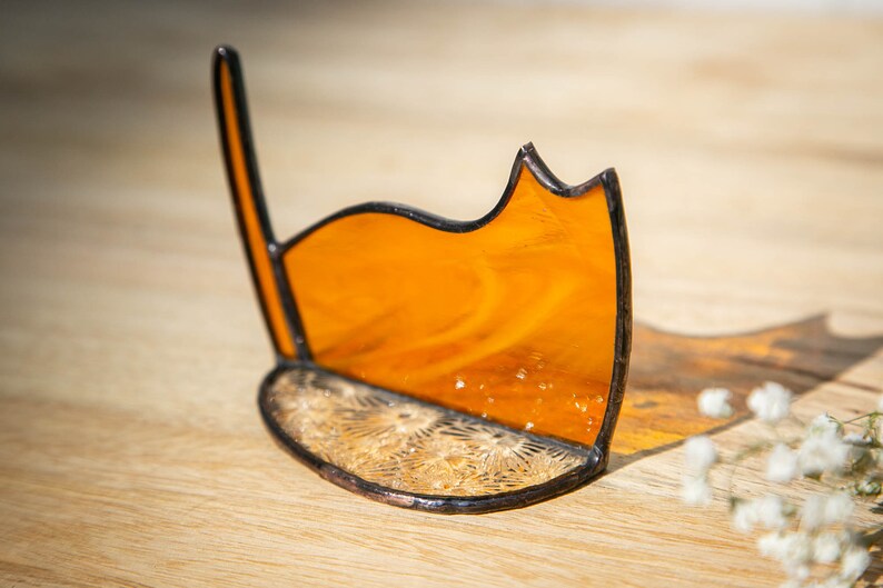 May include: A stained glass cat silhouette ornament featuring a translucent amber-colored body with a dark metal solder frame. The base is a semi-circular piece of clear textured glass with a starburst pattern, joined to the upright cat figure.