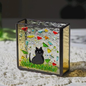 May include: A decorative glass box with a black cat silhouette, colourful accents, and a gold-toned frame. The box features a whimsical design with vibrant red, green, yellow, and blue glass pieces, creating a playful and artistic aesthetic. The cat sits on a patch of green.
