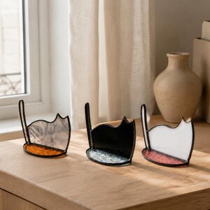 May include: Three stained glass cat-shaped ornaments with dark metal soldering. The left cat is translucent gray with an orange base, the middle is solid black with a clear textured base, and the right is solid white with a pink textured base.