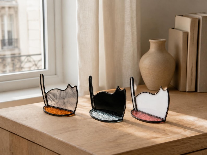May include: Three stained glass cat-shaped ornaments with dark metal soldering. The left cat is translucent gray with an orange base, the middle is solid black with a clear textured base, and the right is solid white with a pink textured base.